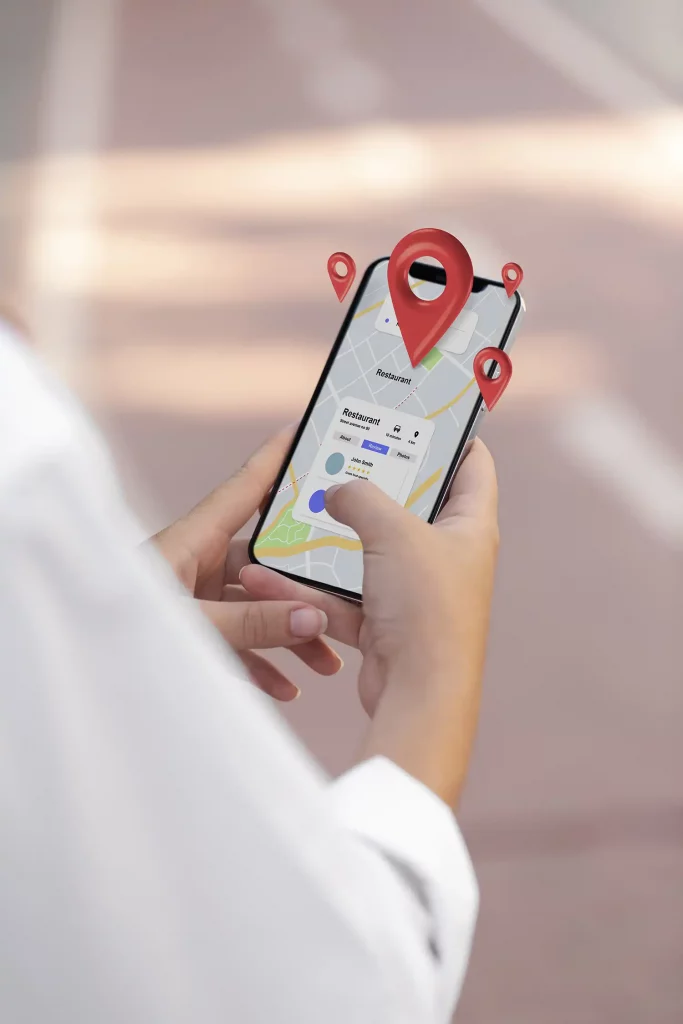 Eine Person hält ein Smartphone in der Hand, auf dessen Display eine Navigationskarte mit einem Restaurant-Profil und Bewertungen zu sehen ist. Über dem Bildschirm schweben rote 3D-Standort-Pins, die symbolisch für die lokale Auffindbarkeit und Präsenz in Google Maps stehen.
